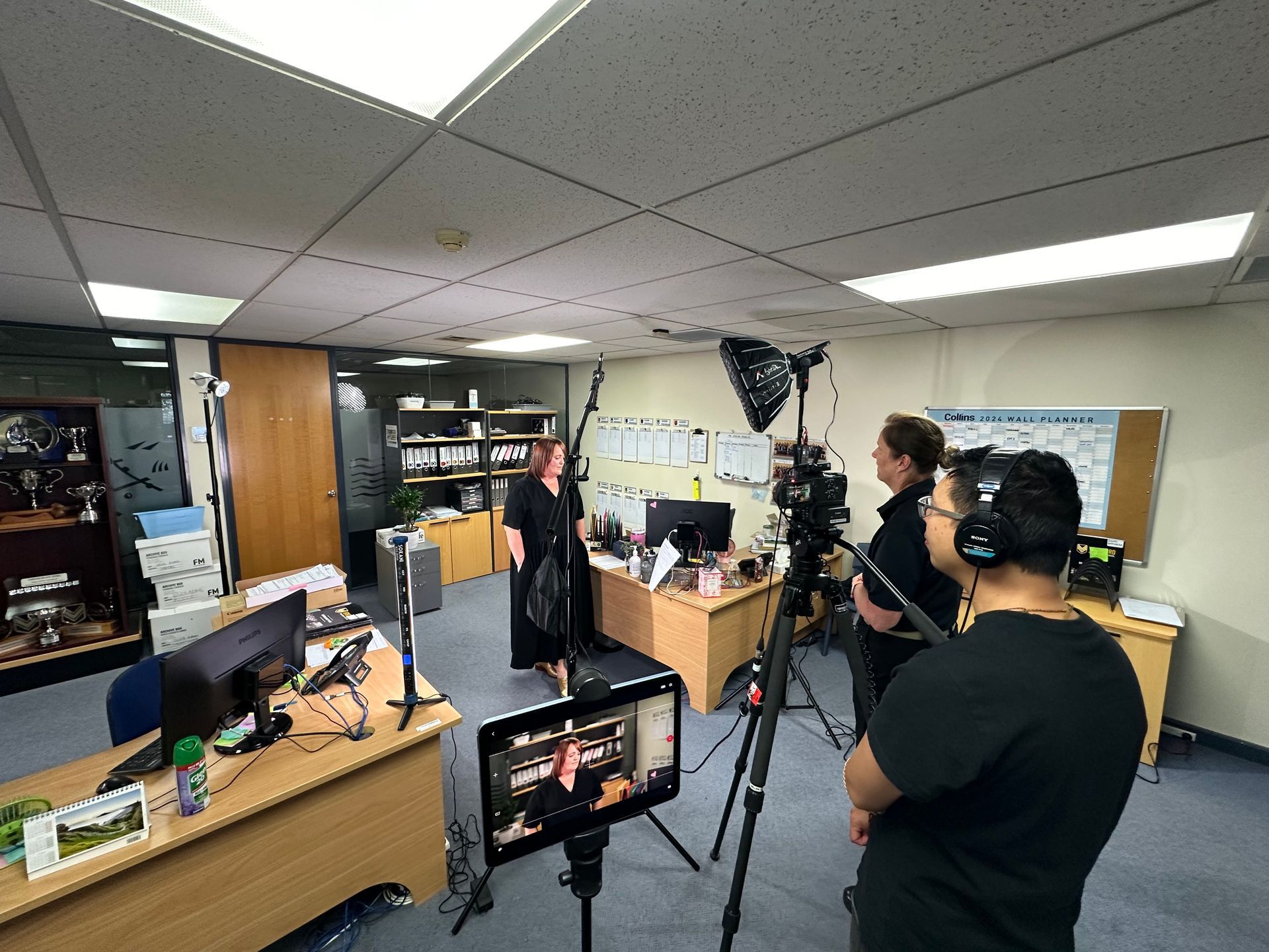 Film crew filming a woman in an office. Camera on tripod, lights, monitor showing the subject.