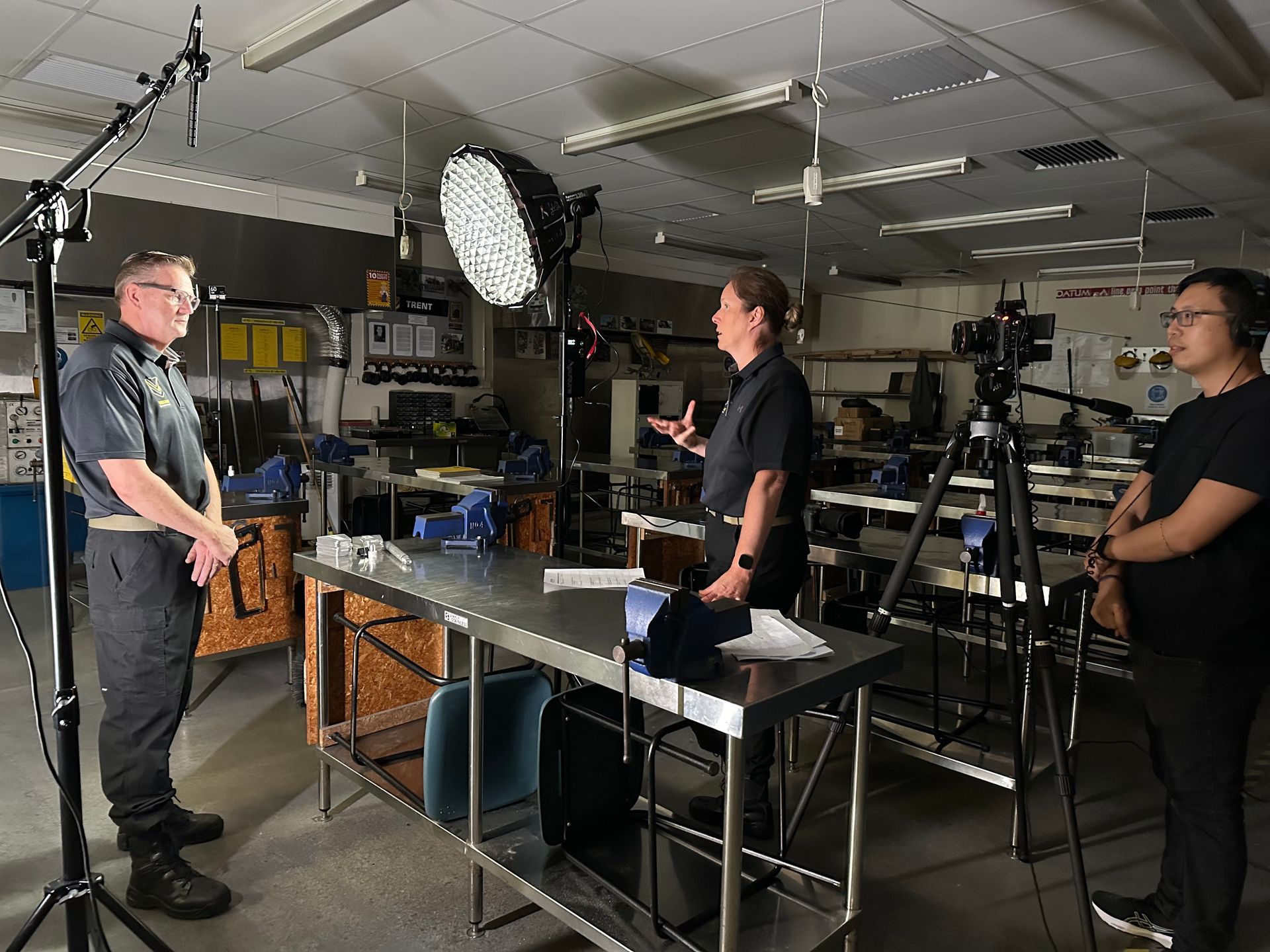 Two people are being filmed in a workshop. A person speaks while the other listens, a camera operator stands by.