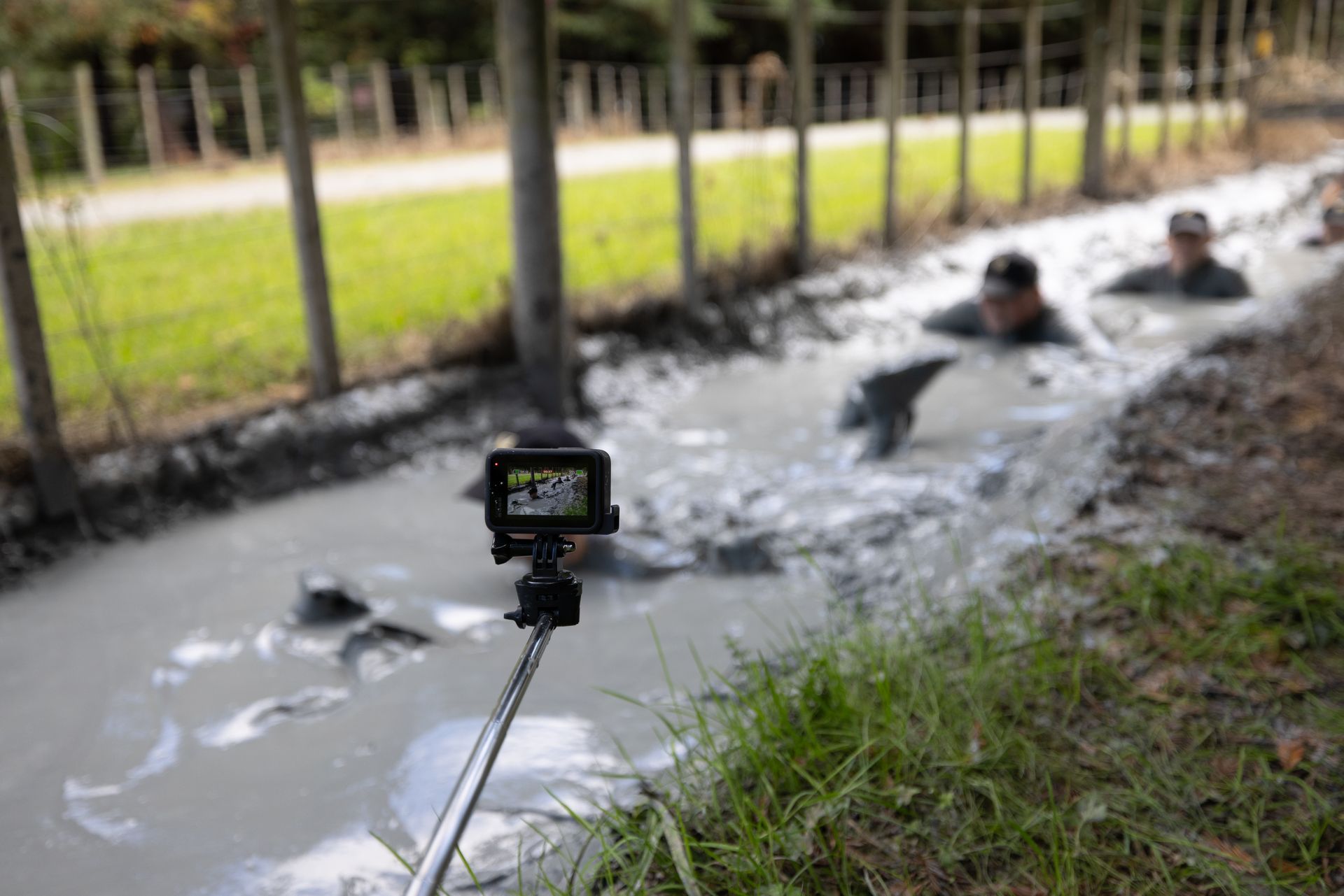 People in a muddy trench, taking a selfie with a GoPro. They are crawling through the mud near a fence.