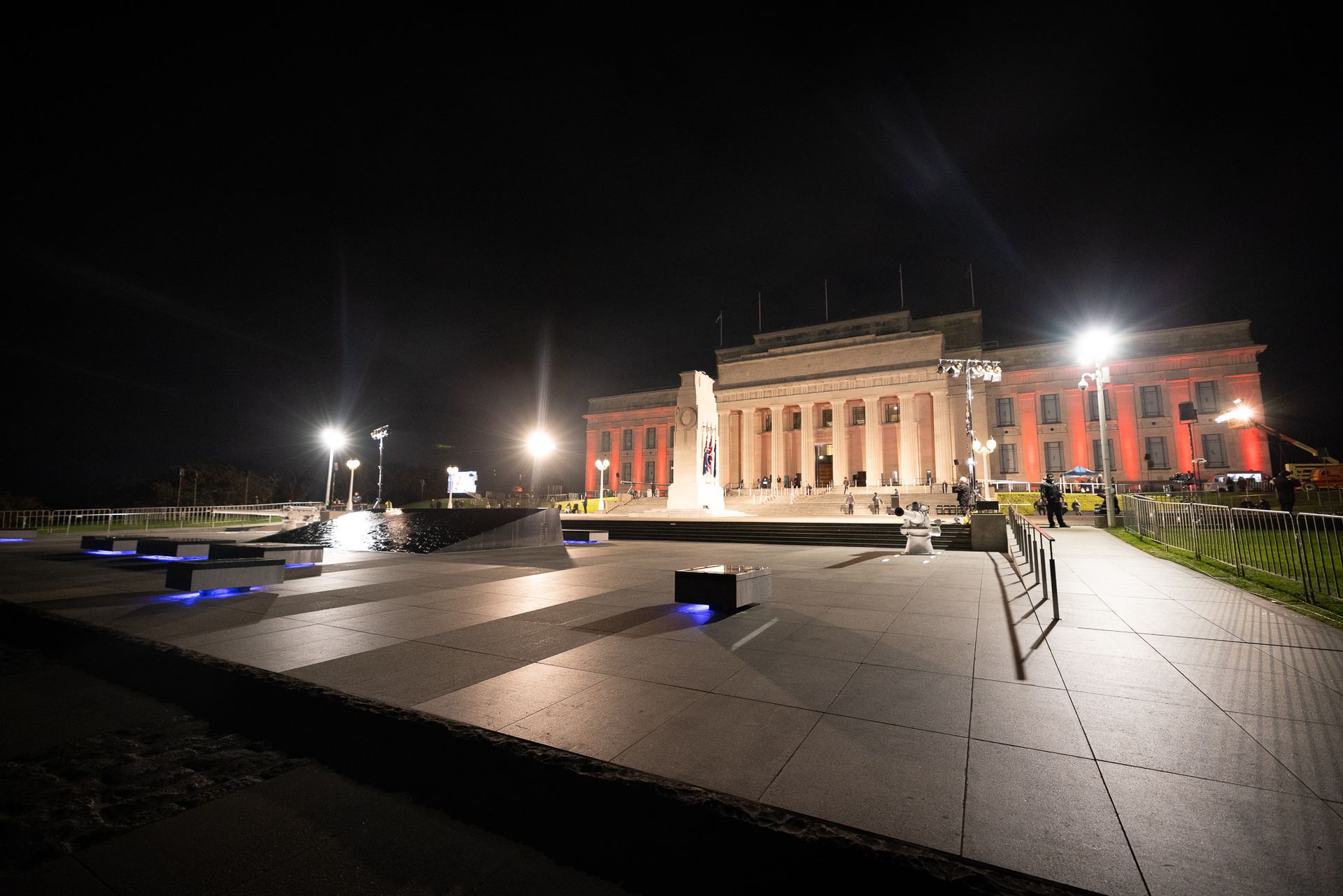 Night view of a lit building with columns, in a plaza with benches and lights.