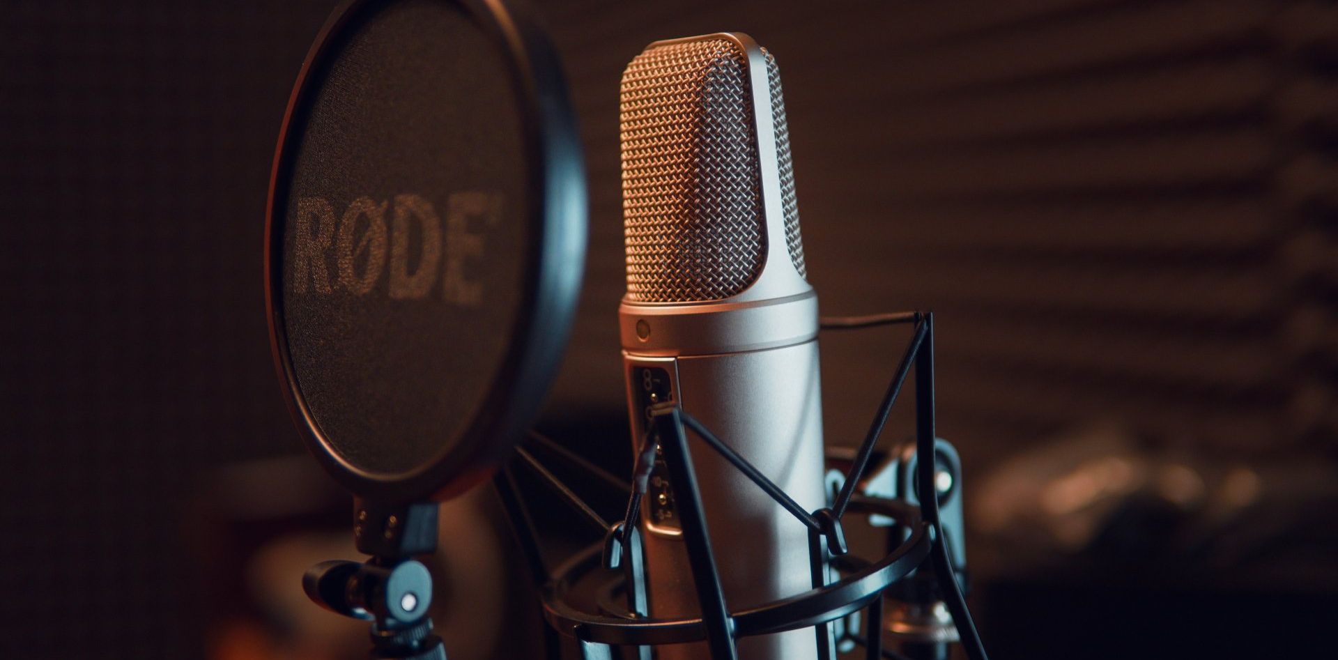 Close-up of a studio microphone and pop filter, with the RODE logo visible, in a dimly lit recording studio.