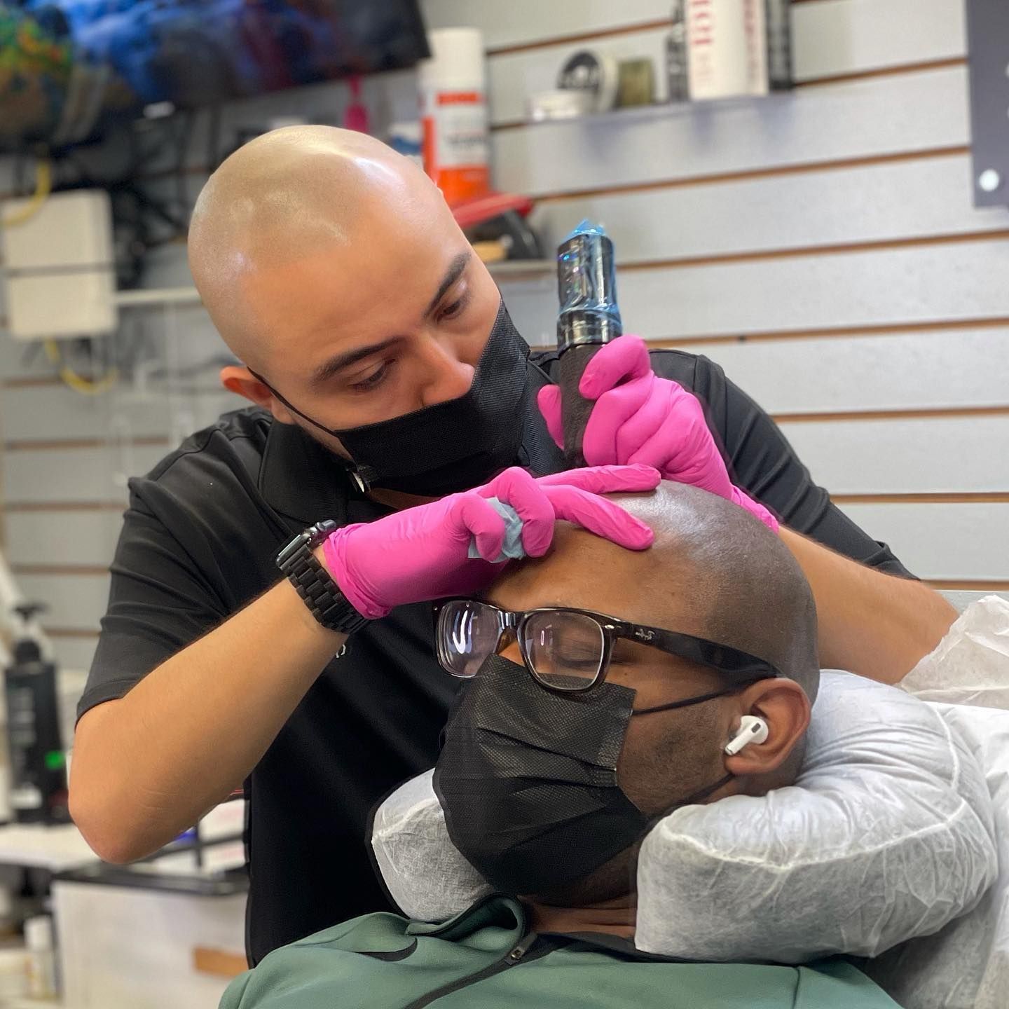A man wearing a mask and pink gloves is getting a tattoo on his head.