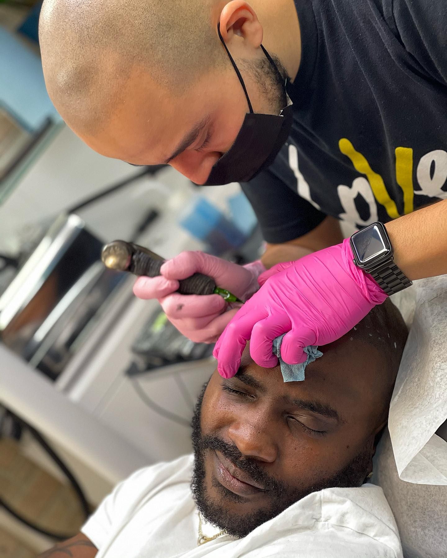 A man wearing a mask and pink gloves is getting a tattoo on his forehead