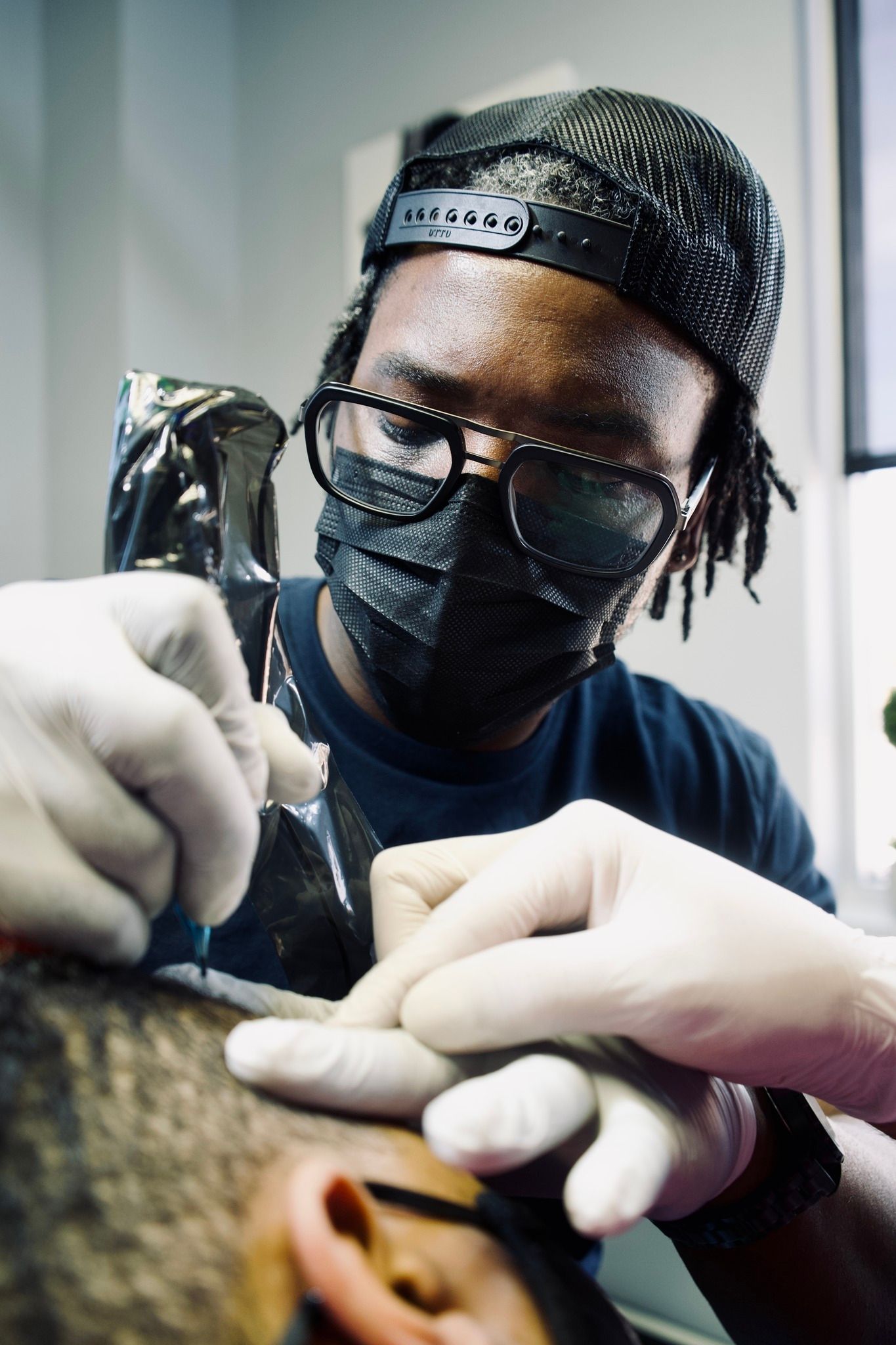A man wearing a mask and gloves is getting a tattoo on another man 's head.