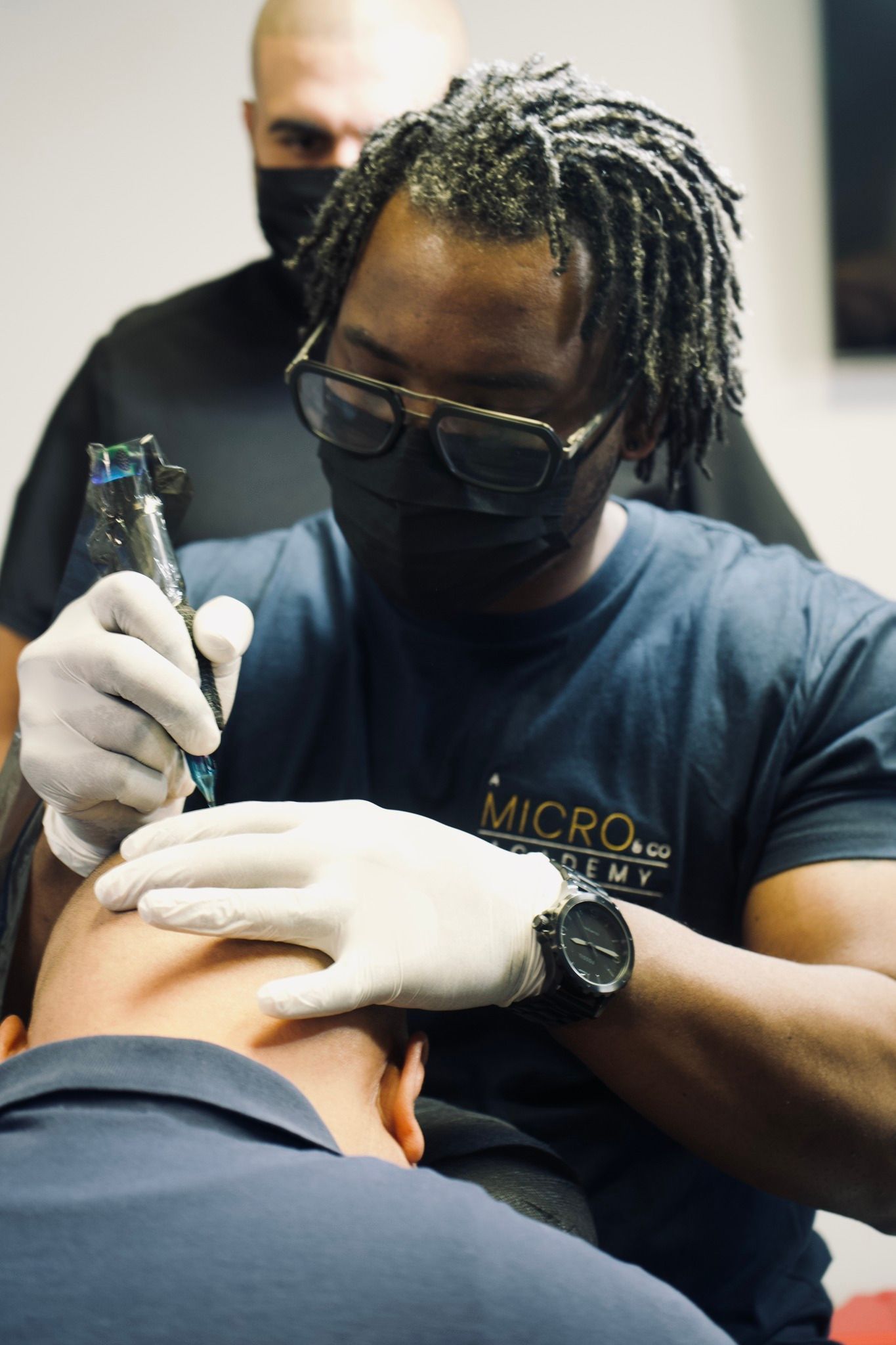 A man wearing a mask and gloves is getting a tattoo on another man 's head.