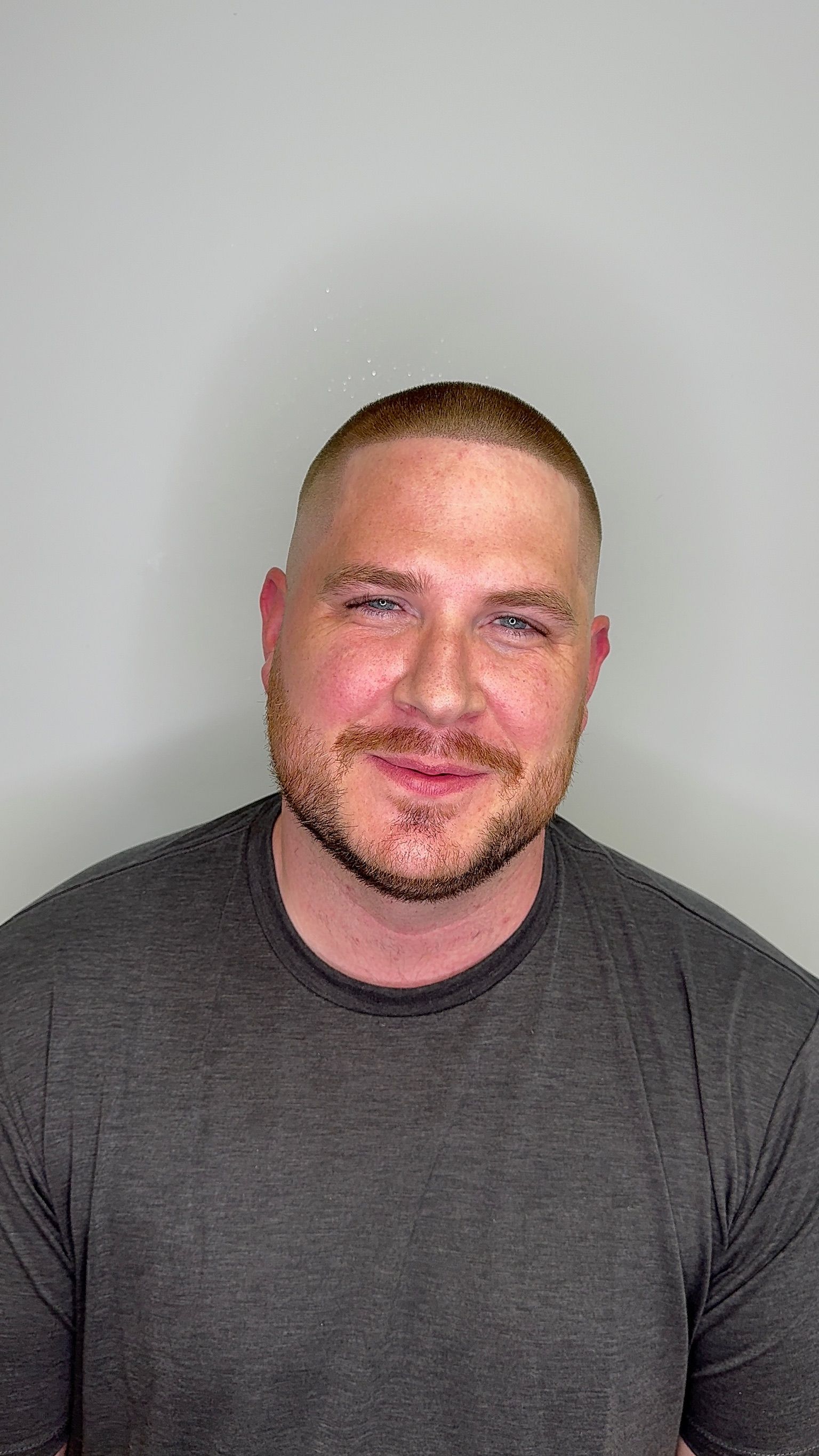 A man with a beard is wearing a grey shirt and smiling.