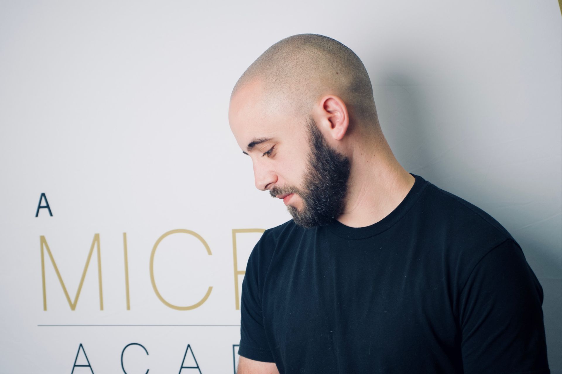 A bald man with a beard is standing in front of a sign that says a mice academy.