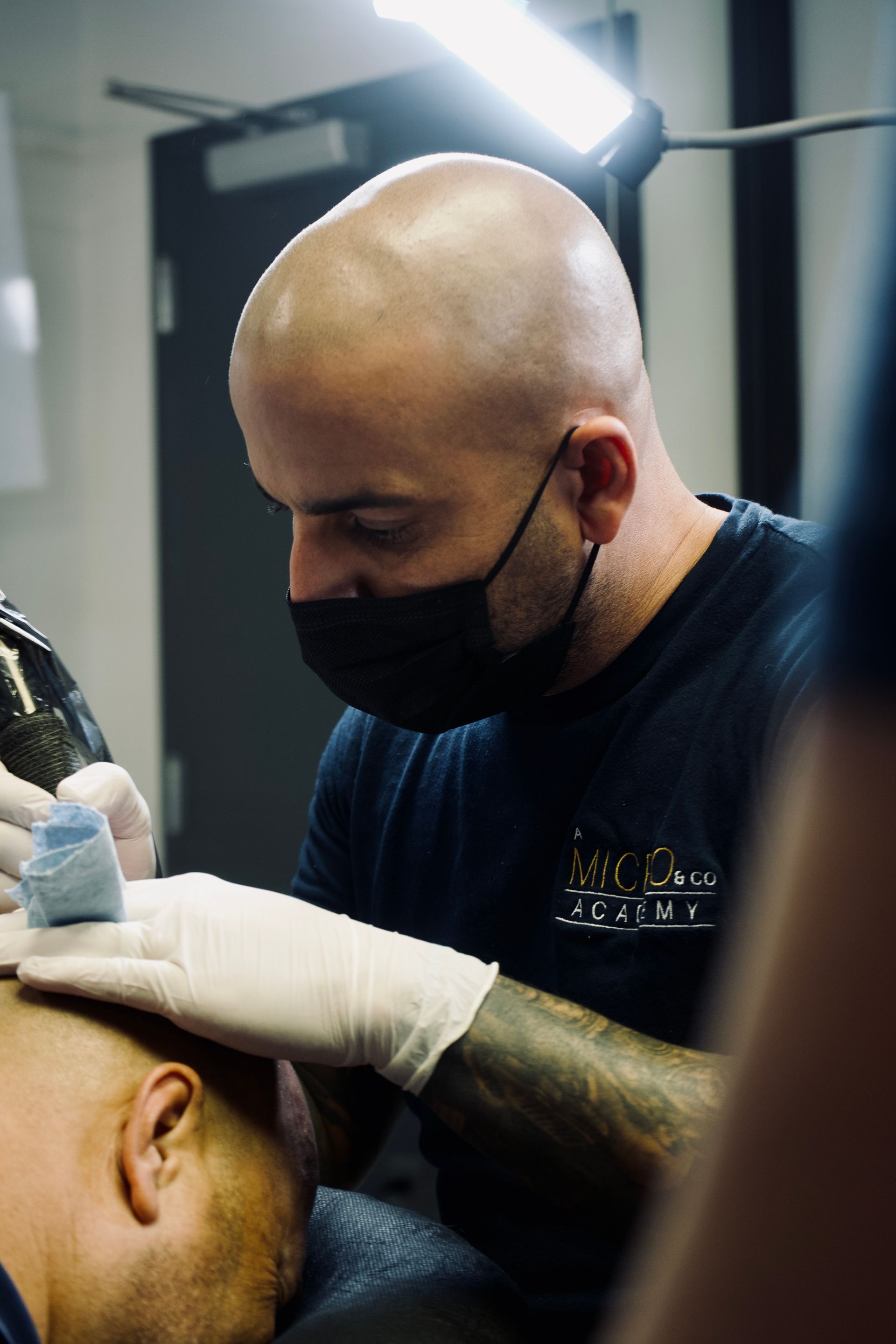 A man wearing a mask and gloves is getting a tattoo on another man 's head.