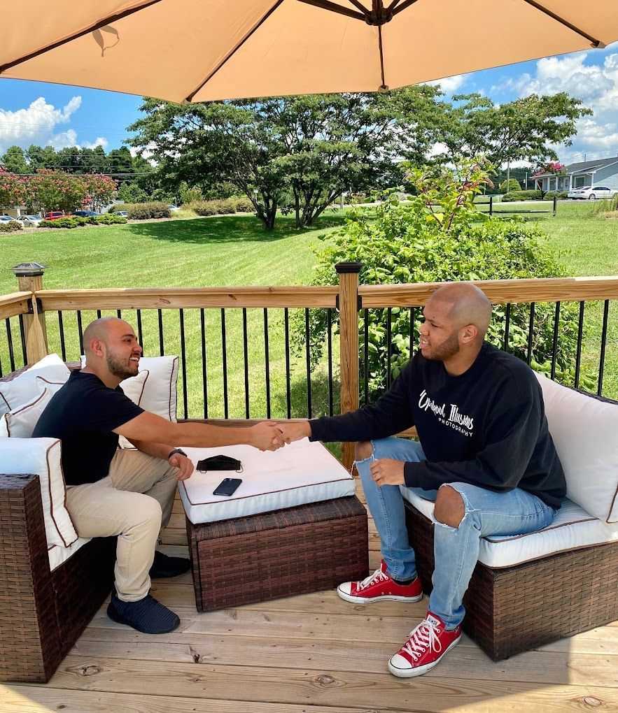 Two men are shaking hands on a deck under an umbrella.