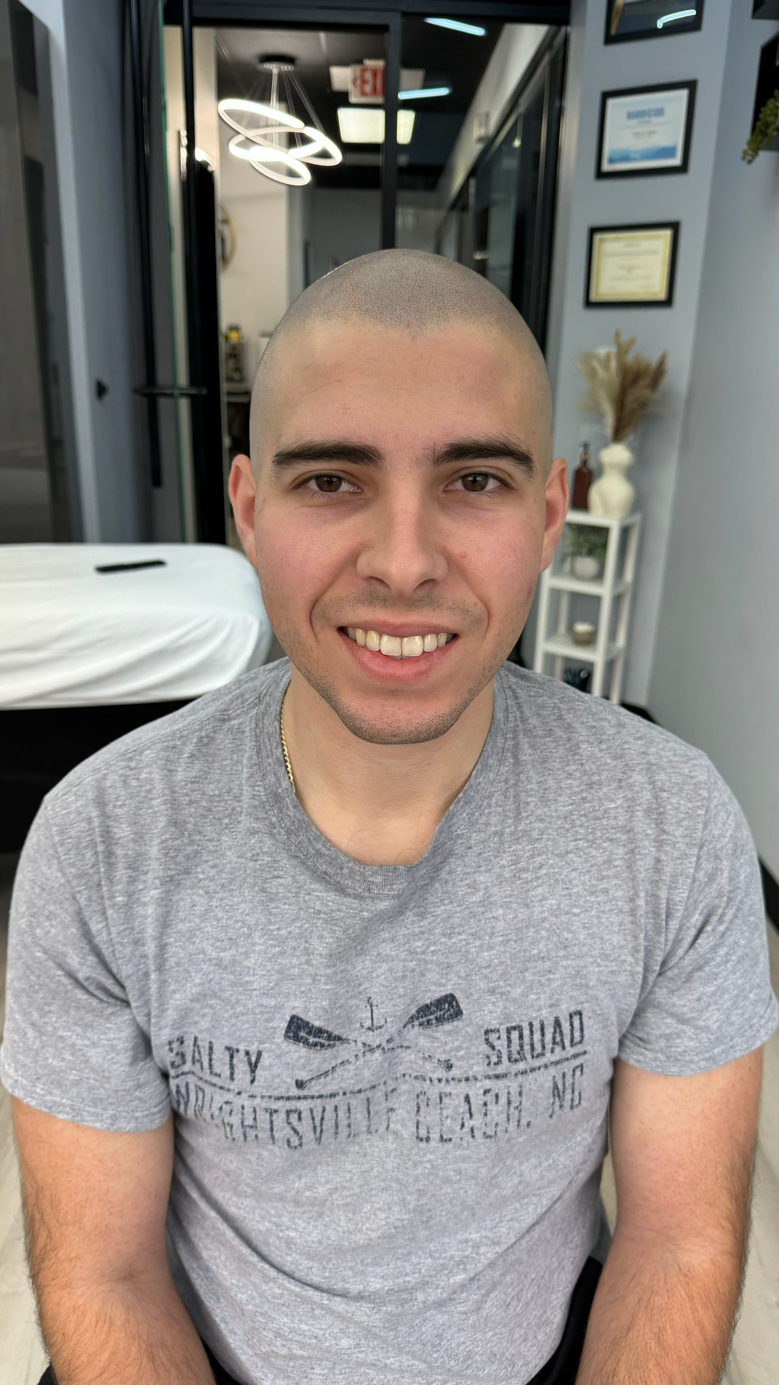 A man with a shaved head is wearing a gray shirt and smiling.