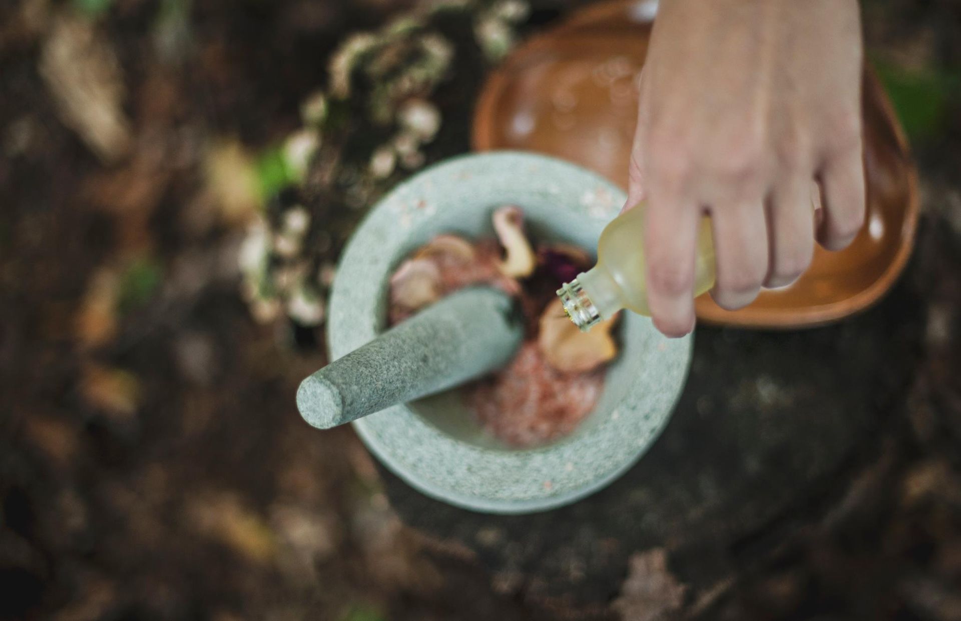 Person pours liquid from a bottle into a mortar containing ingredients, set outdoors on a stump with leaves.