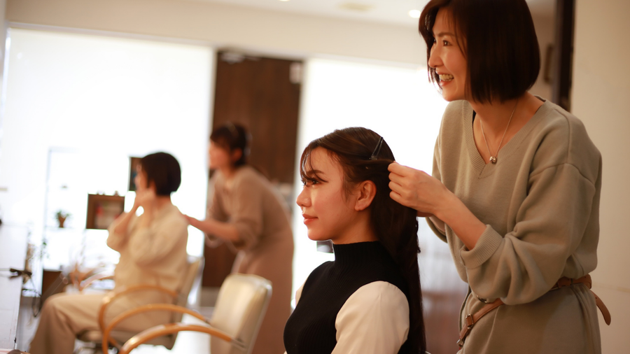 Woman getting her hair styled at a salon; stylist braiding hair, other clients in background.