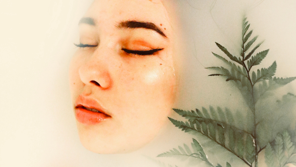 Woman with closed eyes in milky water, a fern next to her face.