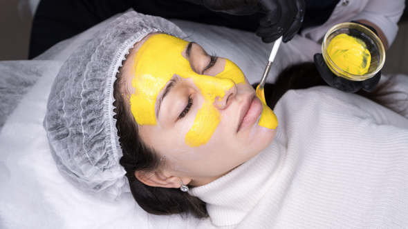 A person receiving a yellow facial mask treatment at a spa; the mask is being applied with a spatula.