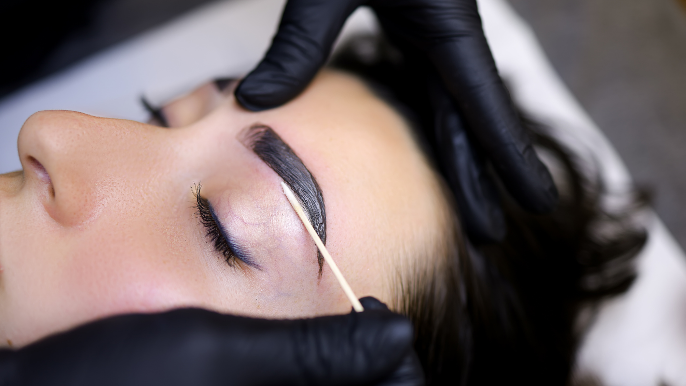 Eyebrow tinting: A person's brow being treated with dark dye by gloved hands, in a spa setting.