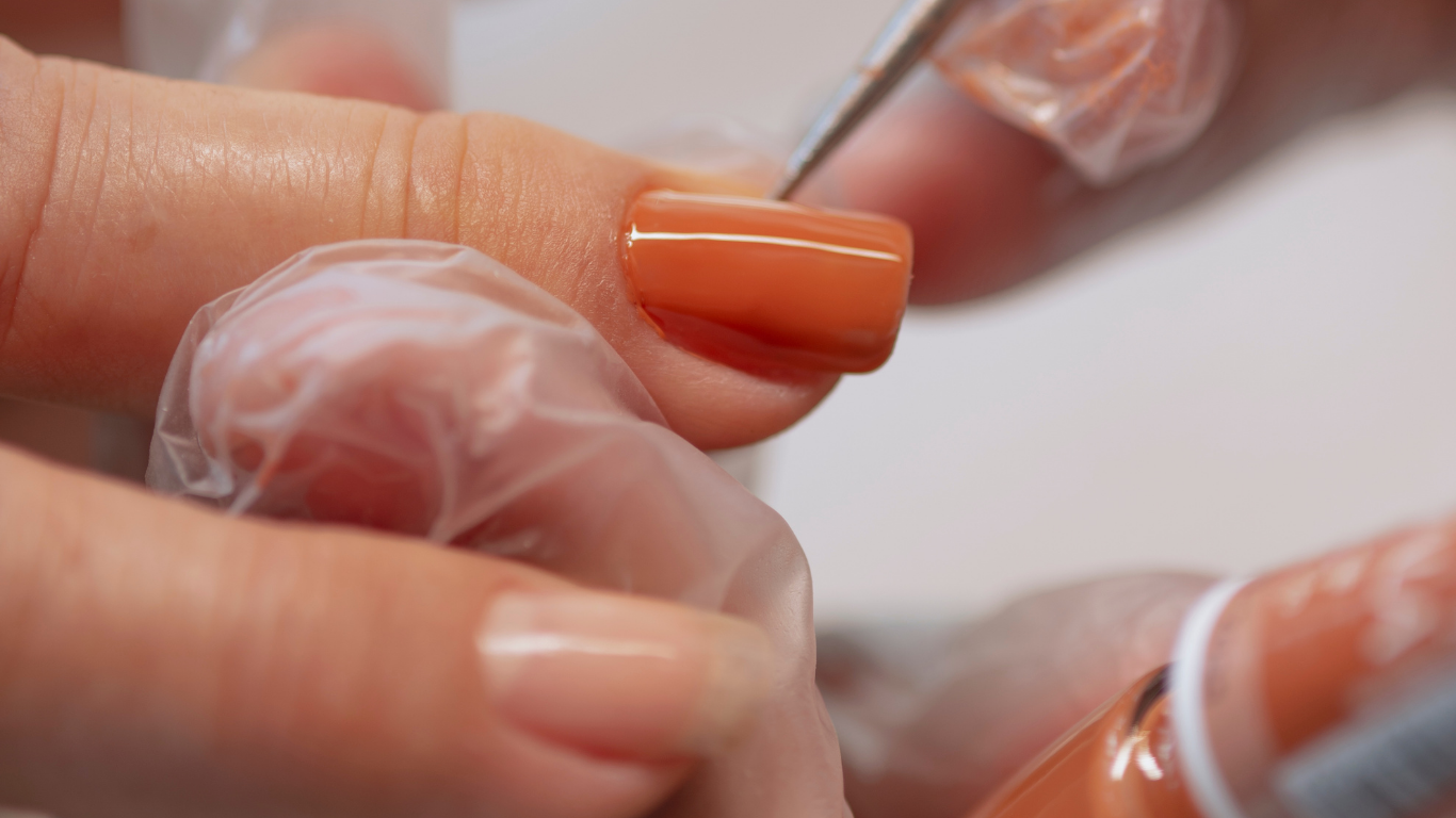 Person applying orange nail polish to a finger in a salon, close-up with gloved hands.