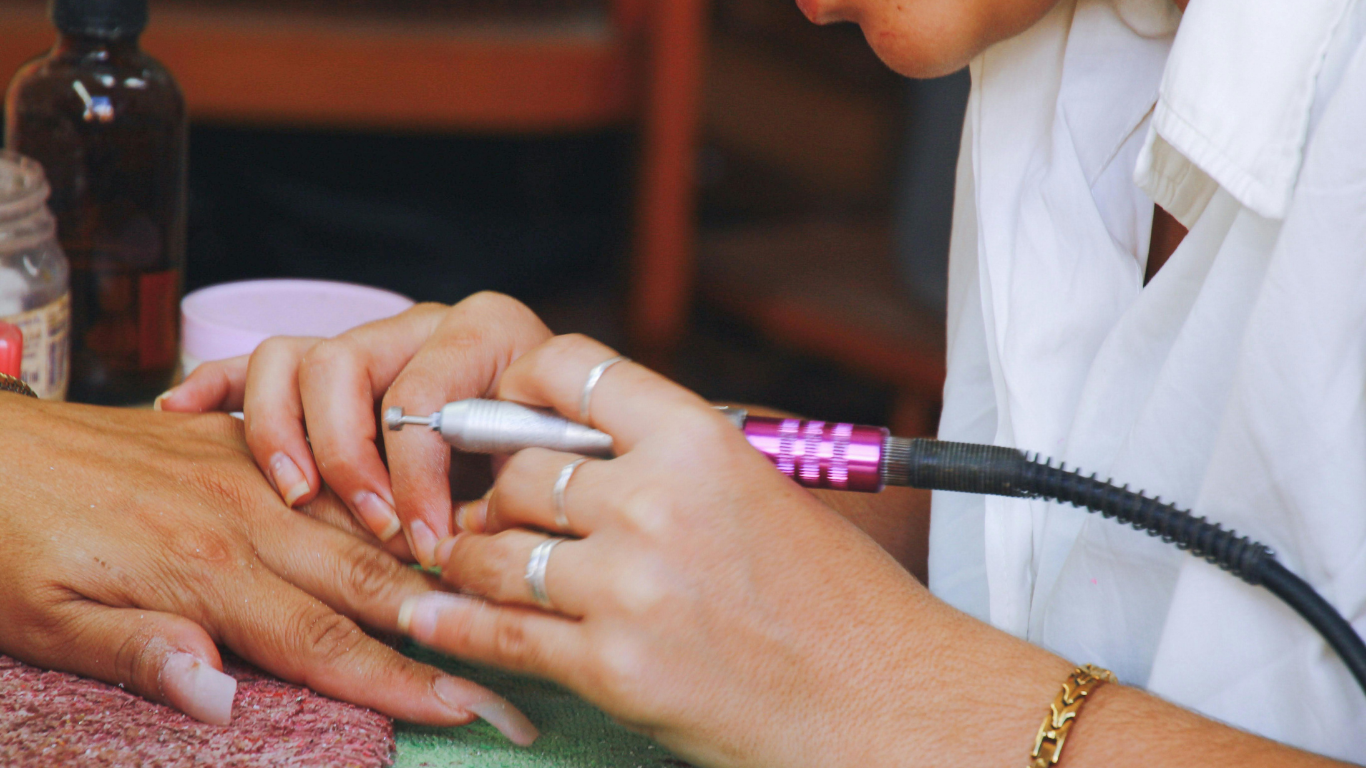 Person using a nail drill on a hand, in a well-lit setting. The operator holds the hand while working on the nails.