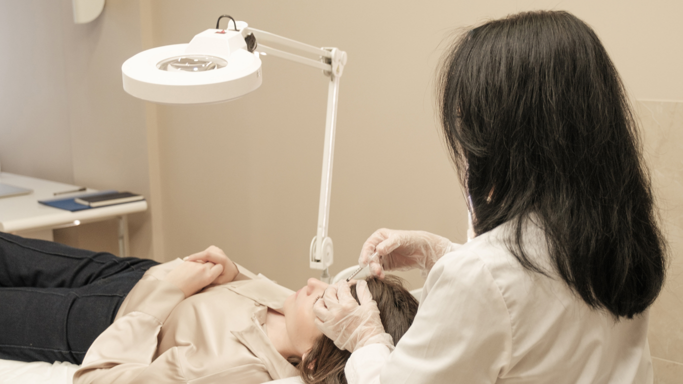 Woman receiving cosmetic treatment on her forehead. Practitioner, in gloves, holds syringe under magnifying lamp.