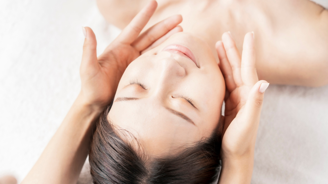 Person receiving a facial massage, hands cupping face and neck, eyes closed, on white.