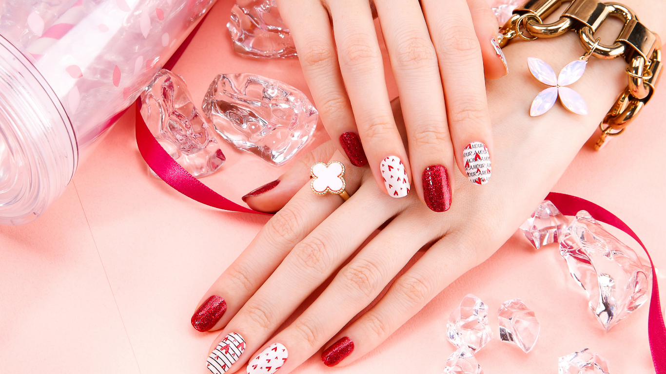 Hands with red and white patterned nail polish, gold bracelet, clover ring, pink background with jewels and ribbon.