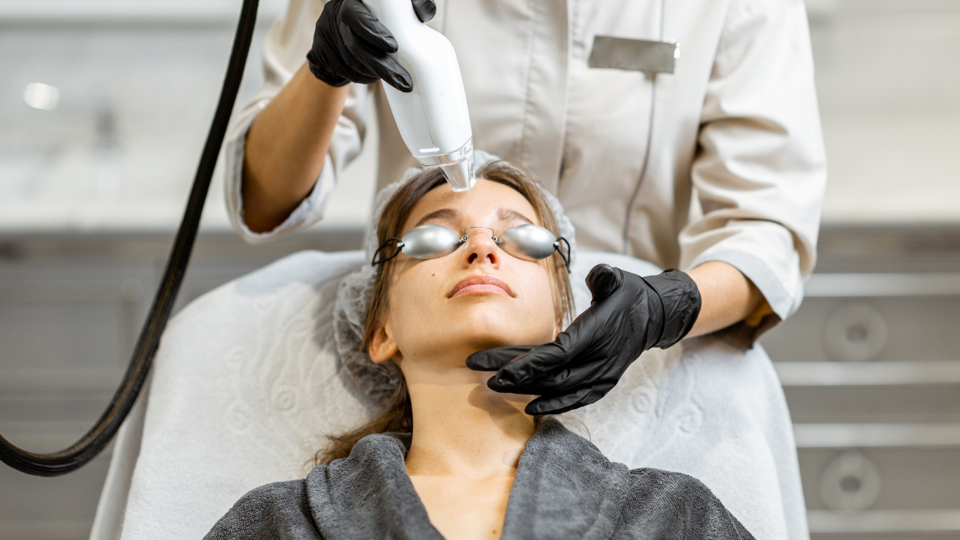 Woman receiving facial laser treatment at a clinic, wearing protective eyewear and robe.