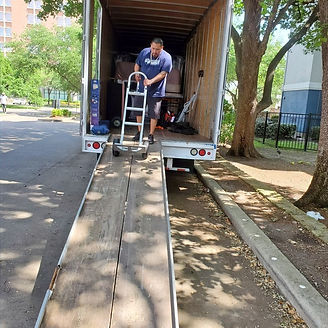 Man loading a moving truck with a hand truck on a wooden ramp outside.