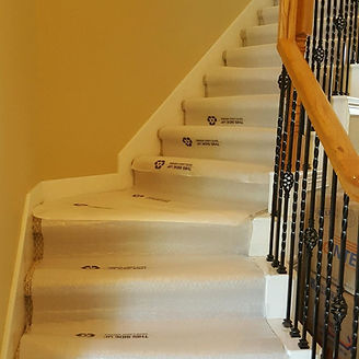 Staircase with carpet protectors, white risers, wooden handrail, black wrought iron balusters, tan wall.