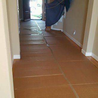 Hallway with brown paper floor covering, beige walls, and a view of an open door to the outside.