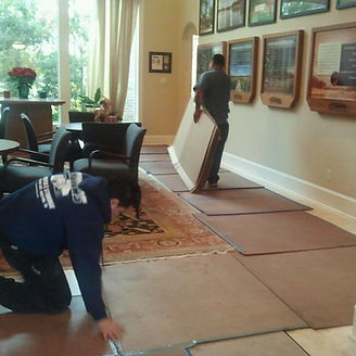 Two men moving furniture, laying protective cardboard on a tile floor in a living room.