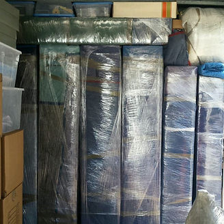 Stacked, wrapped items in a storage unit: blue and brown furniture, boxes, and clear plastic tubs.