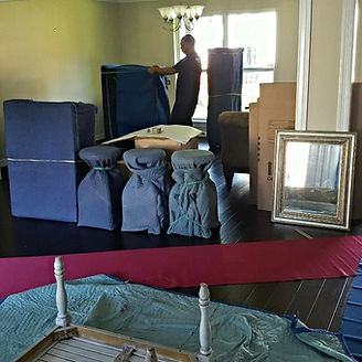 Man packing furniture in a room. Blue-covered items, boxes, mirror, table. Red cloth and tarp on floor.