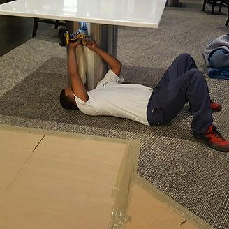 Person lying on floor, using a power drill to assemble a table.