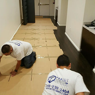 Two people installing flooring in a hallway. One kneels, the other works near dark wood.