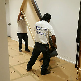 Two people carrying a large rectangular object through a hallway with protective floor covering.