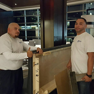 Two men holding a large wooden panel indoors, thumbs up and smiling, with cityscape in the background.