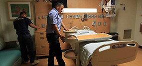 Two men in a hospital room by a bed with a tray. One man touches the tray.