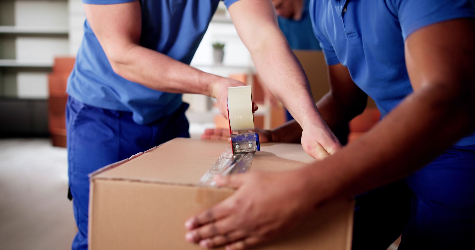 Movers taping up a cardboard box, indoors, using blue tape.