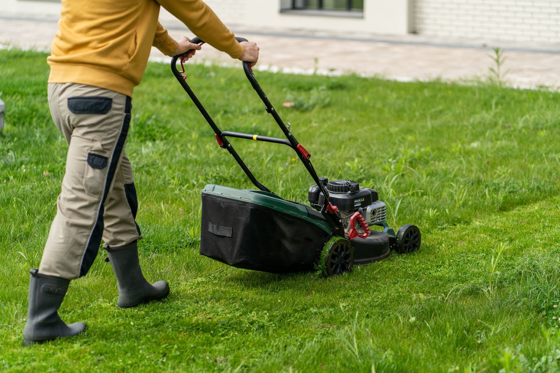 Person mowing a bright green lawn with a black and gold lawnmower near a wooden fence.