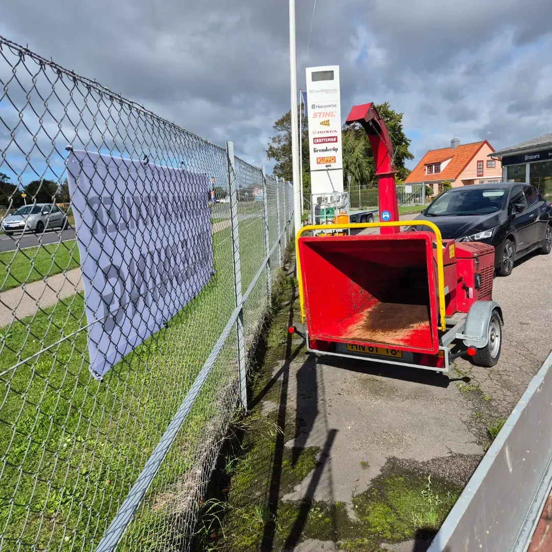 Rød flishugger på en trailer ved siden af et hegn, en vej og en bygning.