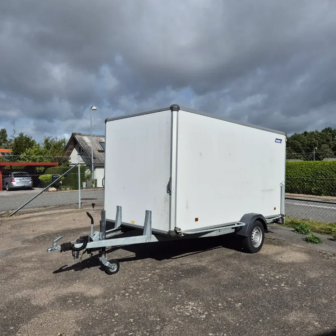 Hvid lukket lasttrailer parkeret udendørs på en grå overflade under en overskyet himmel.