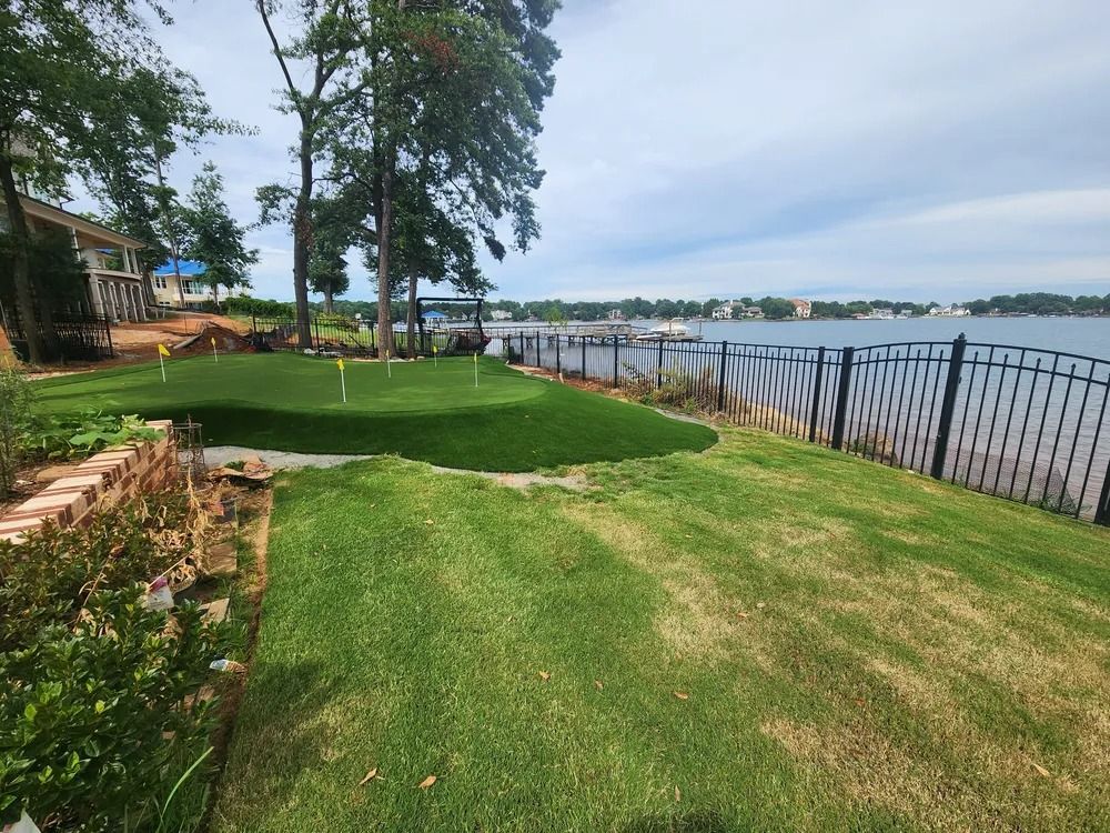 Grassy lakeside yard with trees, a patio, and a black metal fence along the water