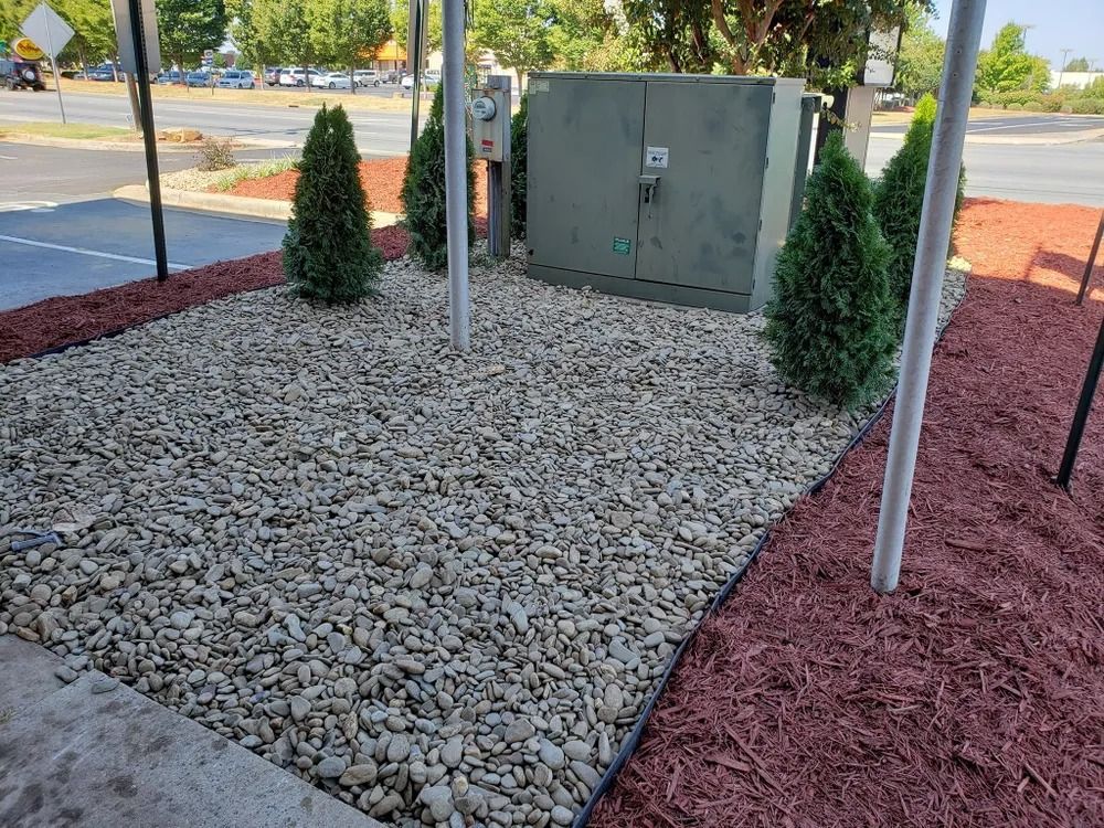 Landscaped utility area with gravel, red mulch, two evergreens, and a gray electrical box near a sidewalk