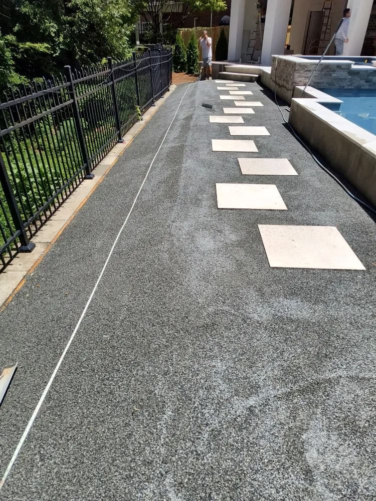 Outdoor walkway with square stepping stones beside a pool and black fence in sunlight