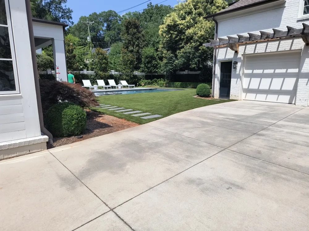 Concrete driveway beside a backyard lawn, pool, and white patio chairs on a sunny day