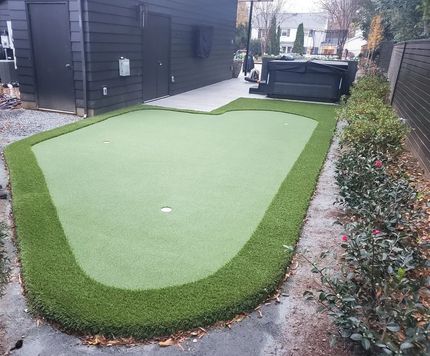 Synthetic putting green in a narrow backyard beside a dark house and patio, bordered by shrubs.