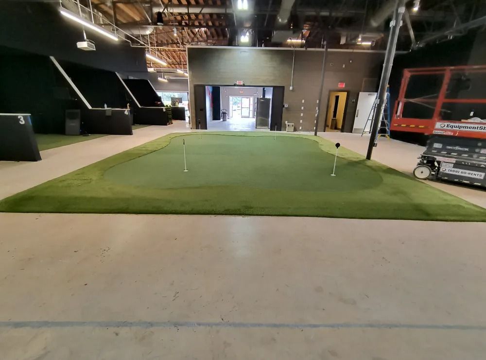 Indoor golf practice area with a putting green, surrounding turf, and construction materials nearby