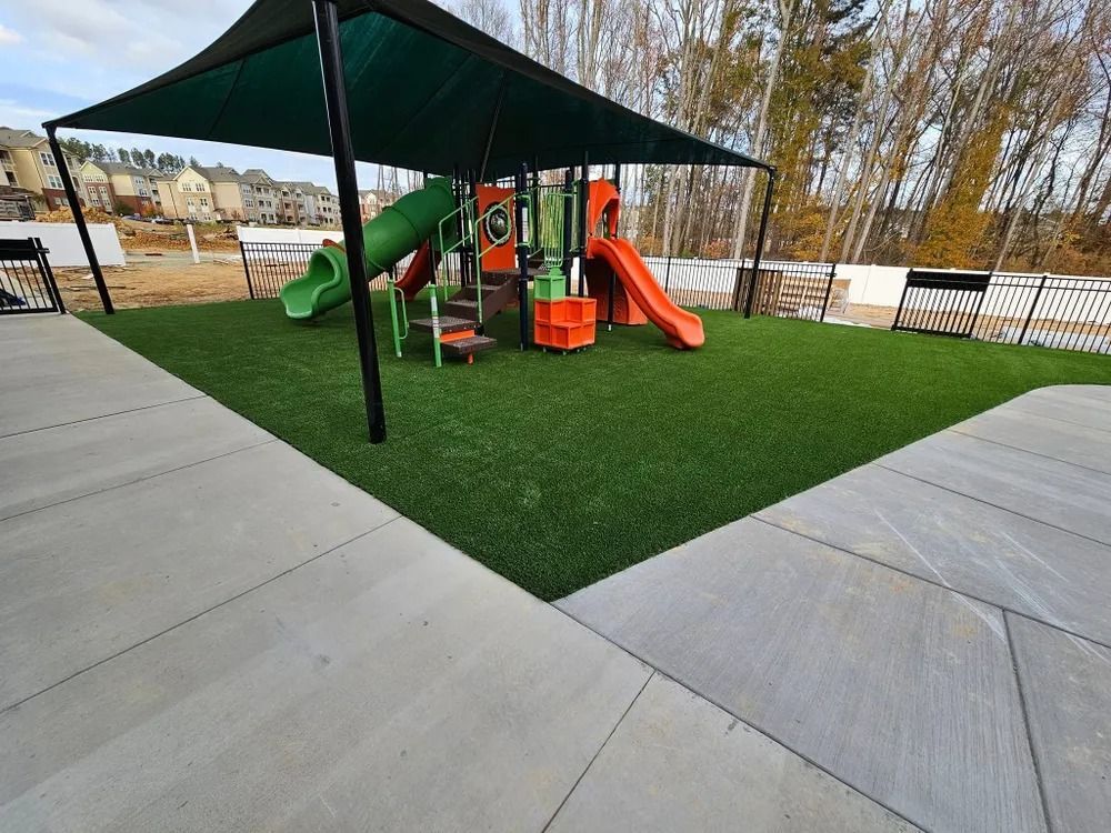 Covered playground with green turf, orange slides, and black shade canopy between concrete walkways.