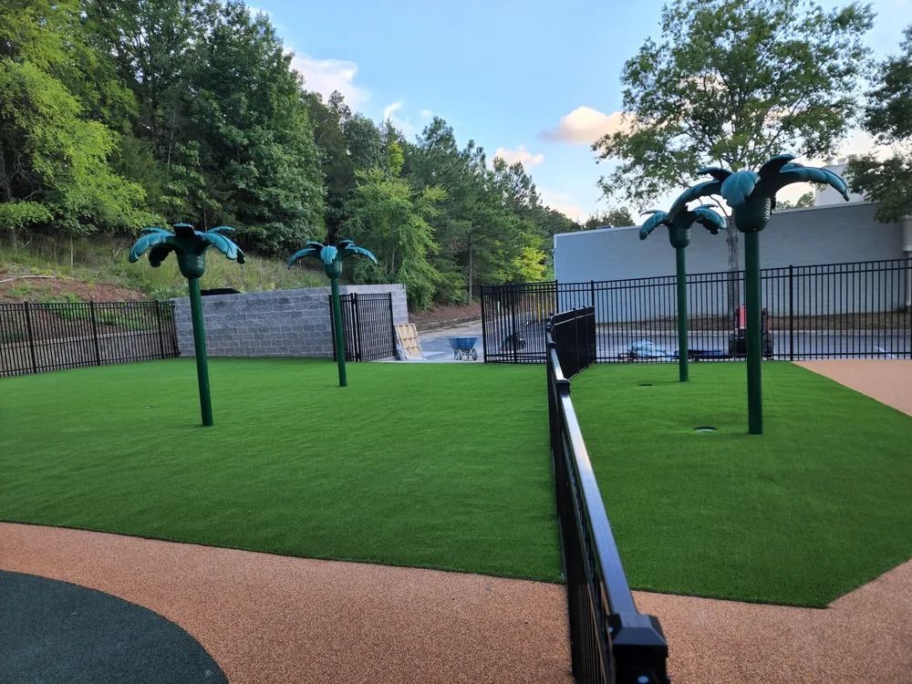 Mini golf putting green with palm tree obstacles, black rail, and a white building by wooded rock walls.