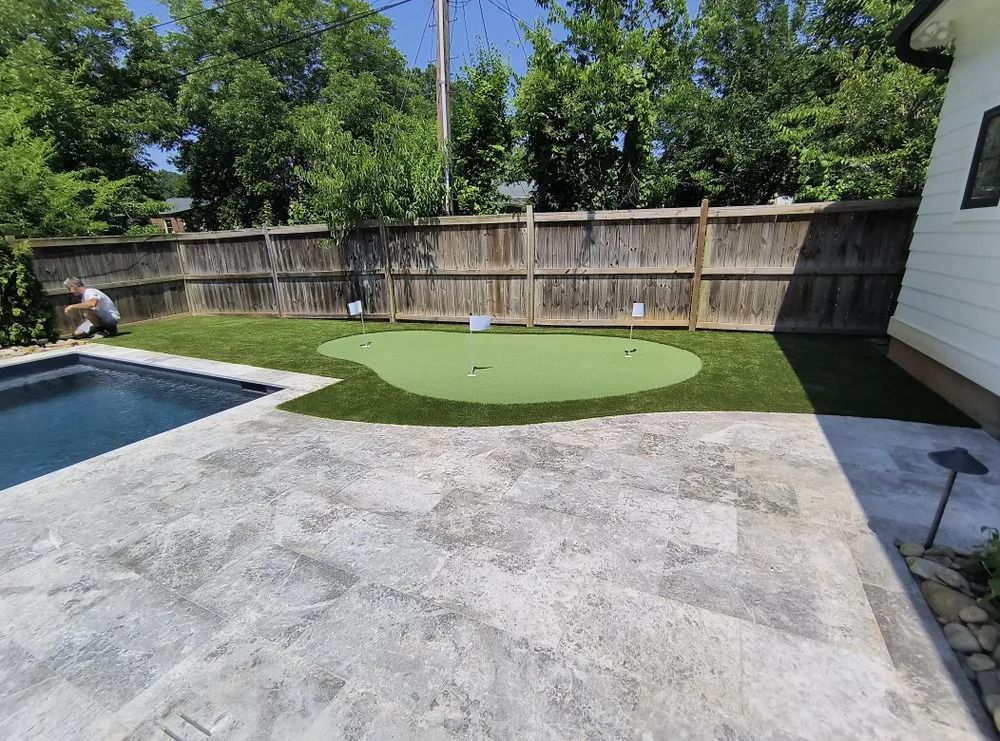Backyard patio with a pool, putting green, concrete deck, and wooden fence under a blue sky