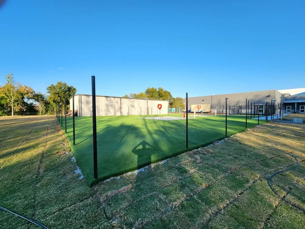 Green fenced batting cage on grassy field beside a building under a blue sky.